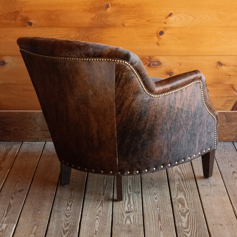 Angled back view of a rustic arm chair with tapered hardwood legs and nailhead trim upholstered in brindle hair-on-hide leather on a wide-plank wooden floor against a rustic wooden wall
