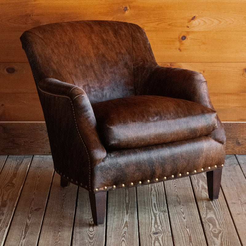 Angled front view of a rustic arm chair with tapered hardwood legs and nailhead trim upholstered in brindle hair-on-hide leather on a wide-plank wooden floor against a rustic wooden wall