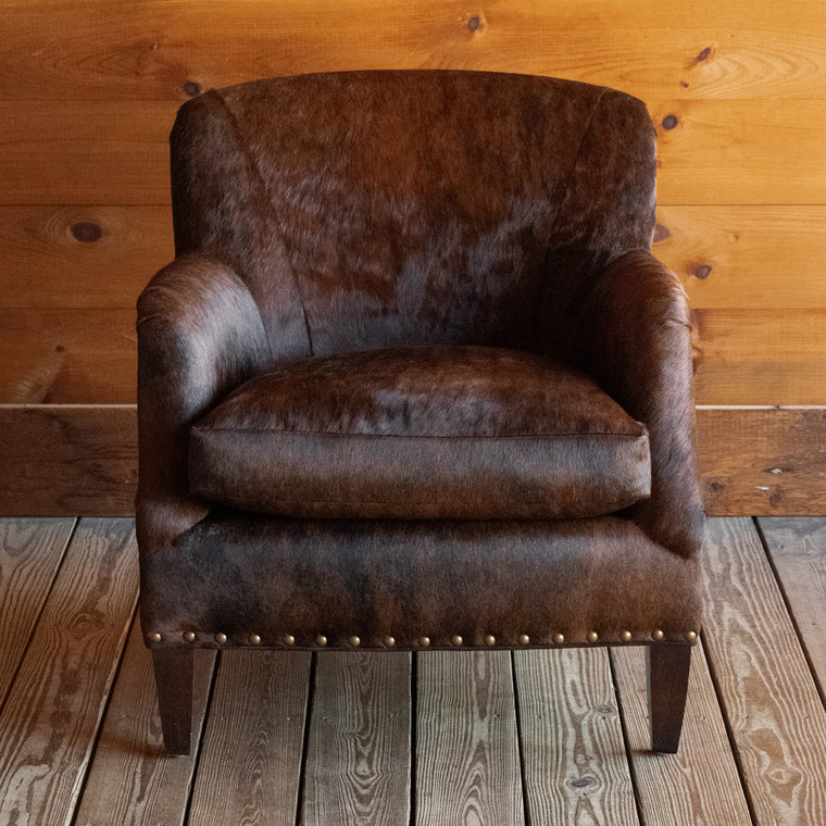 Angled back view of a rustic arm chair with tapered hardwood legs and nailhead trim upholstered in brindle hair-on-hide leather on a wide-plank wooden floor against a rustic wooden wall