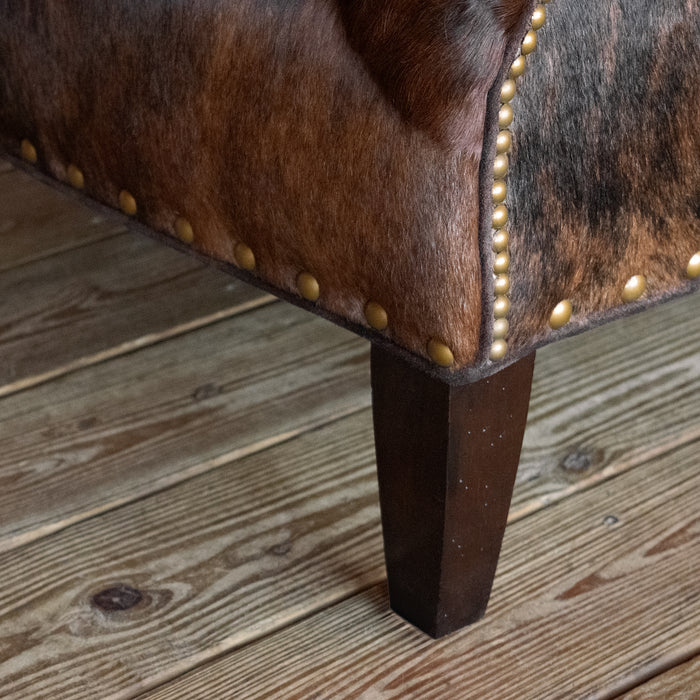 Closeup of the leg and nailhead trim of a rustic arm chair upholstered in brindle hair-on-hide leather on a wide-plank wooden floor