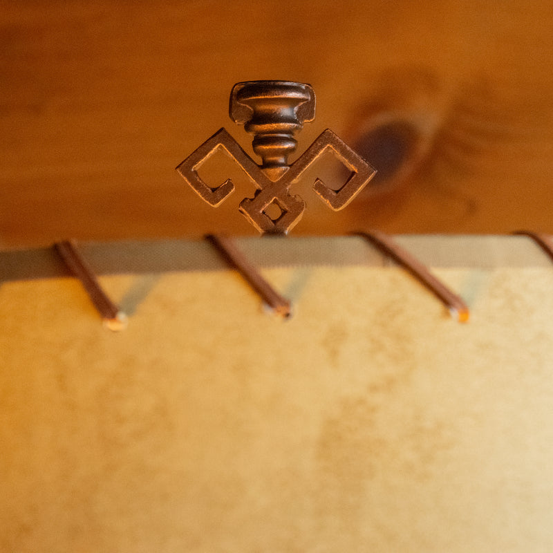 Close-up of the finial of a rustic floor lamp with an oxidized bronze finish and a calfskin shade with leather lacing against a rustic wooden wall