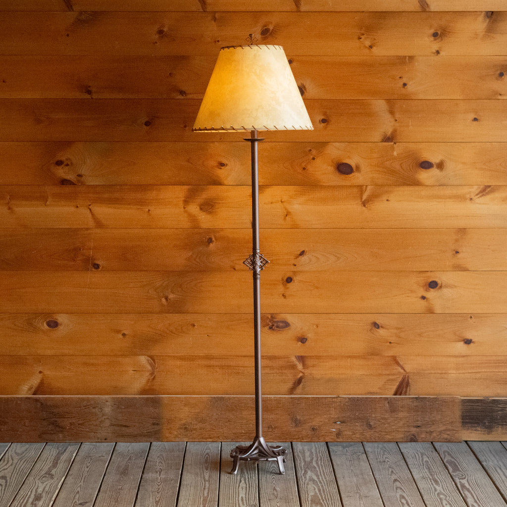 Front view of a rustic floor lamp with an oxidized bronze finish and a calfskin shade with leather lacing on wide-plank wooden floor against a rustic wooden wall