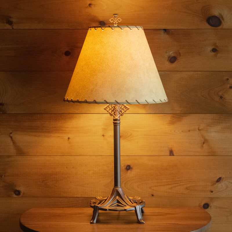 Front view of a rustic table lamp with an oxidized bronze finish and a calfskin shade with leather lacing on a wooden table against a rustic wooden wall