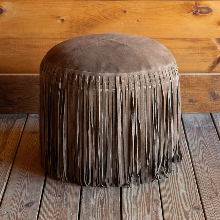 Rustic brown suede leather ottoman with fringe on a wooden floor against a wooden wall