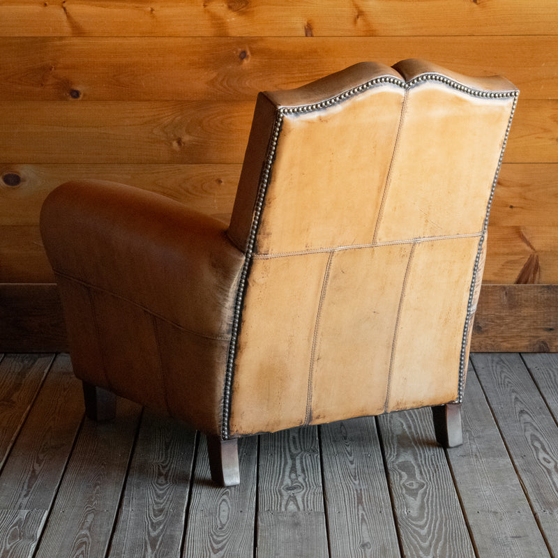 Angled back view of a rustic buffalo leather armchair with a tight back and nailhead trim on a wooden floor against a wooden wall