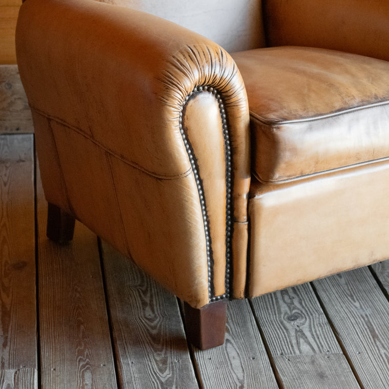 Angled back view of a rustic buffalo leather armchair with a tight back and nailhead trim on a wooden floor against a wooden wall