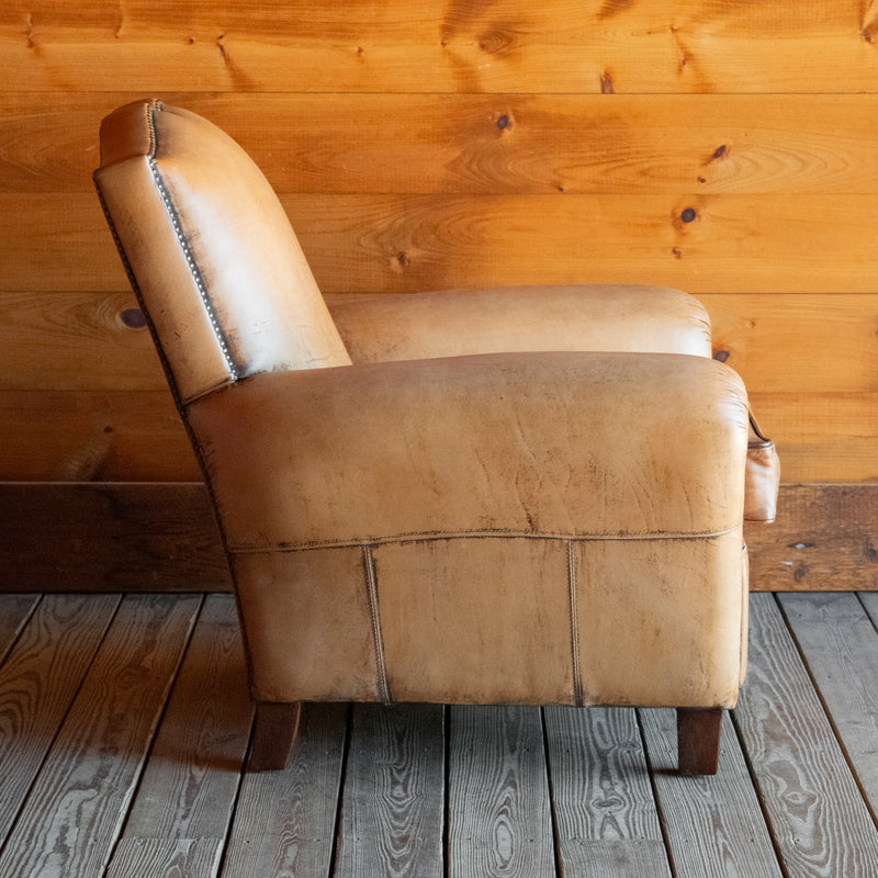 Profile view of a rustic buffalo leather armchair with a tight back and nailhead trim on a wooden floor against a wooden wall