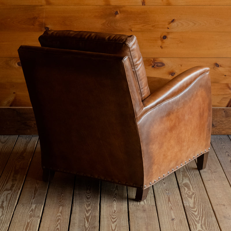 Angled back view of a rustic brown burnished leather chair with nailhead trim and tapered wooden legs on a wide-plank wood floor against a rustic wooden wall