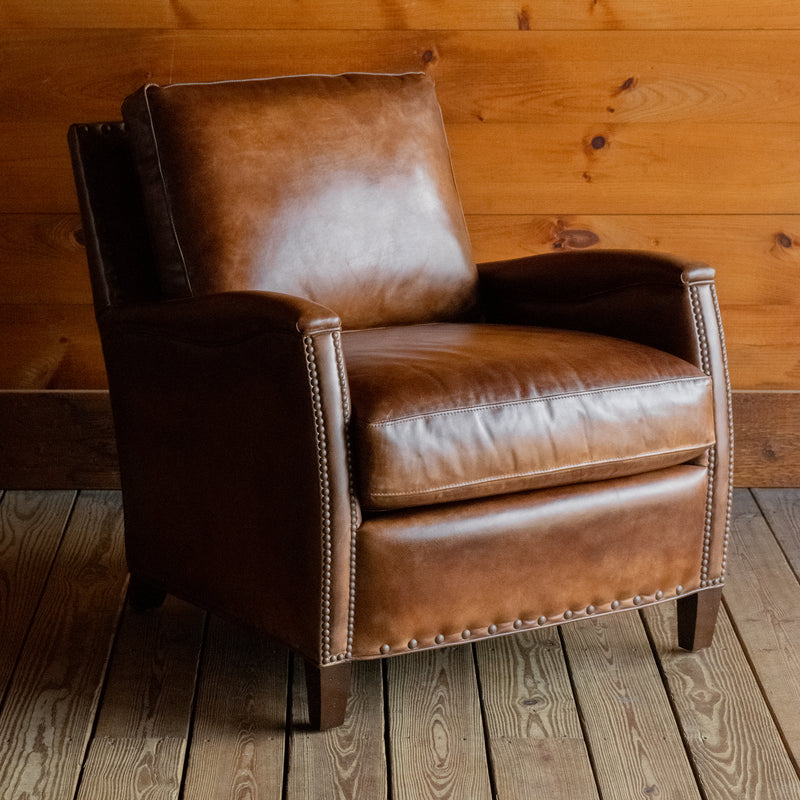 Angled front view of a rustic brown burnished leather chair with nailhead trim and tapered wooden legs on a wide-plank wood floor against a rustic wooden wall