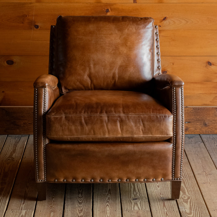 Front view of a rustic brown burnished leather chair with nailhead trim and tapered wooden legs on a wide-plank wood floor against a rustic wooden wall