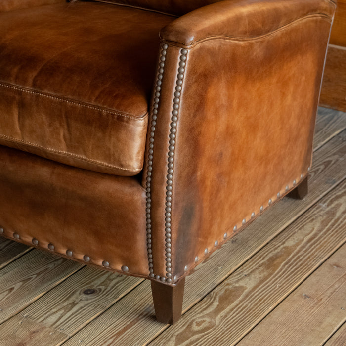 Close-up of the arm and leg of a rustic brown burnished leather chair with nailhead trim and tapered wooden legs on a wide-plank wood floor
