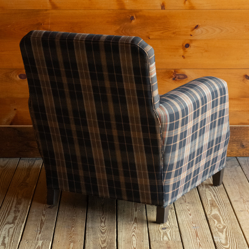 Angled back view of a club chair upholstered in a dark plaid fabric with nailhead trim on a wide-plank wooden floor against a rustic wooden wall