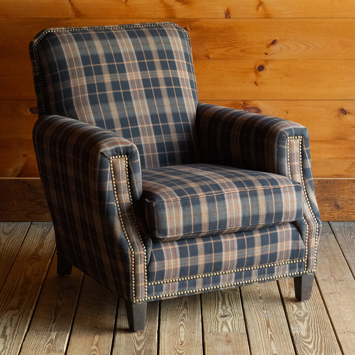 Angled front view of a club chair upholstered in a dark plaid fabric with nailhead trim on a wide-plank wooden floor against a rustic wooden wall