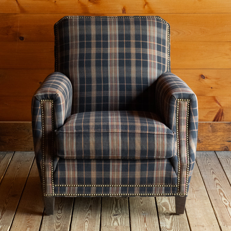 Angled back view of a club chair upholstered in a dark plaid fabric with nailhead trim on a wide-plank wooden floor against a rustic wooden wall