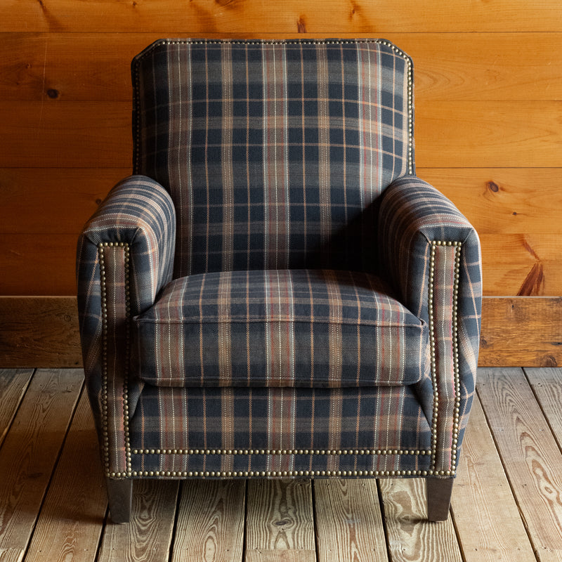 Front view of a club chair upholstered in a dark plaid fabric with nailhead trim on a wide-plank wooden floor against a rustic wooden wall