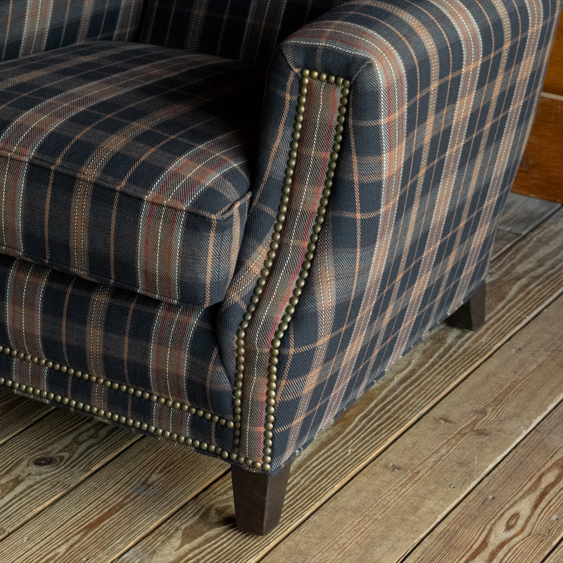 Close-up of the arm of club chair upholstered in a dark plaid fabric with nailhead trim on a wide-plank wooden floor