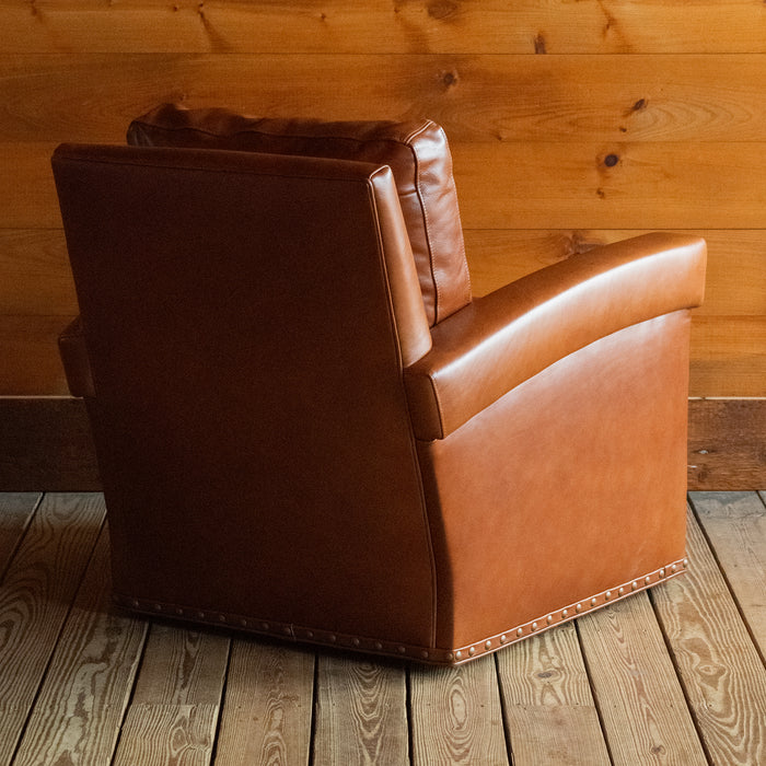 Angled back view of a rustic cognac leather swivel chair with nailhead and gimp trim on a wide-plank wooden floor against a rustic wooden wall
