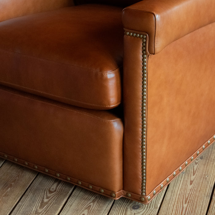 Close-up of the arm, seat cushion, nailhead detail, and gimp trim of a rustic cognac leather swivel chair on a wide-plank wooden floor