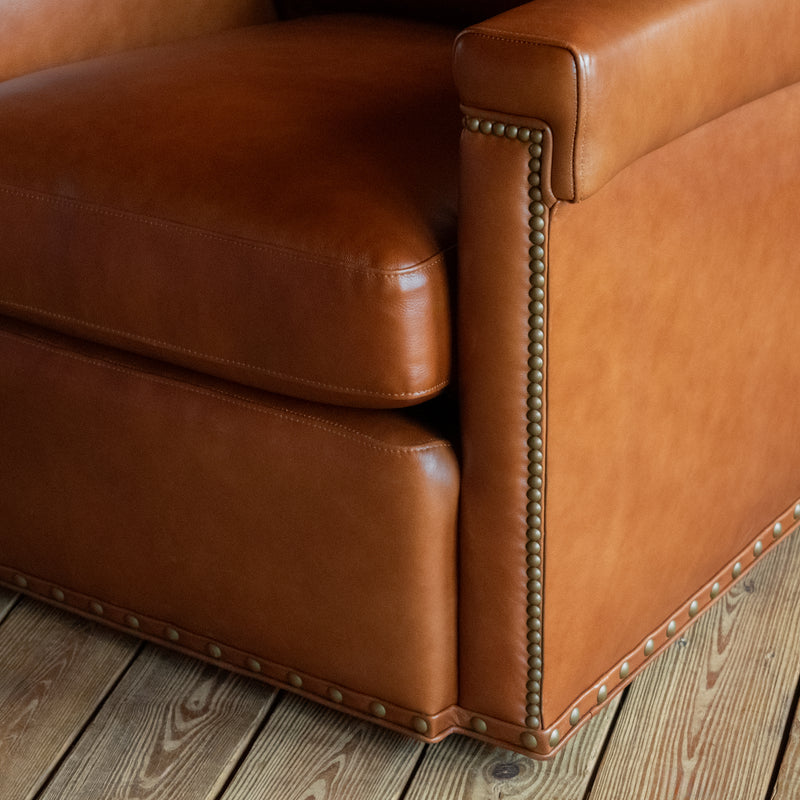 Close-up of the arm, seat cushion, nailhead detail, and gimp trim of a rustic cognac leather swivel chair on a wide-plank wooden floor