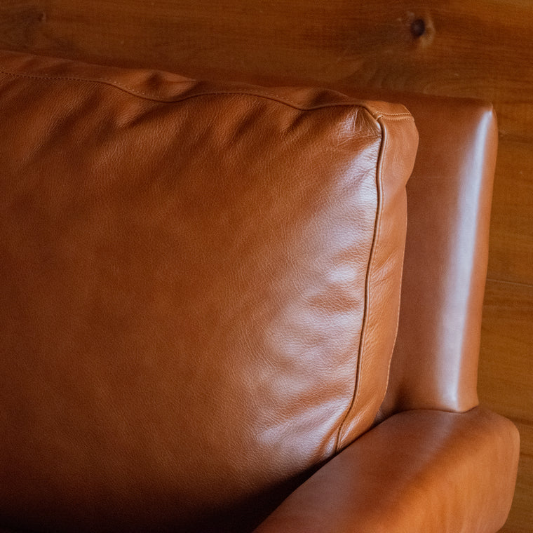 Close-up of the back corner of a rustic cognac leather swivel chair against a rustic wooden wall