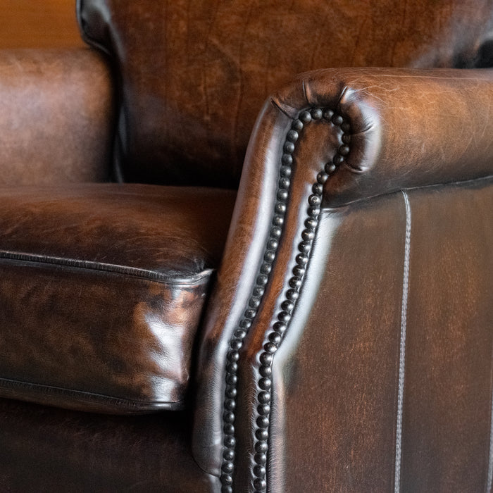 Close-up of the arm of a rustic dark brown leather chair against a wooden wall 
