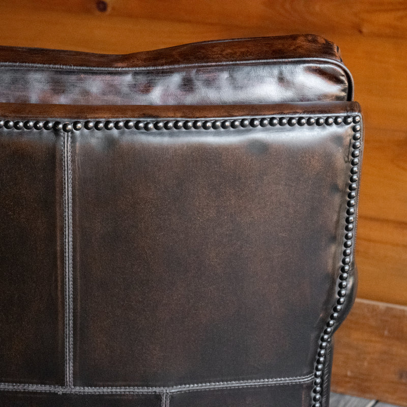 Close-up of the back of a rustic dark brown leather chair against a wooden wall 