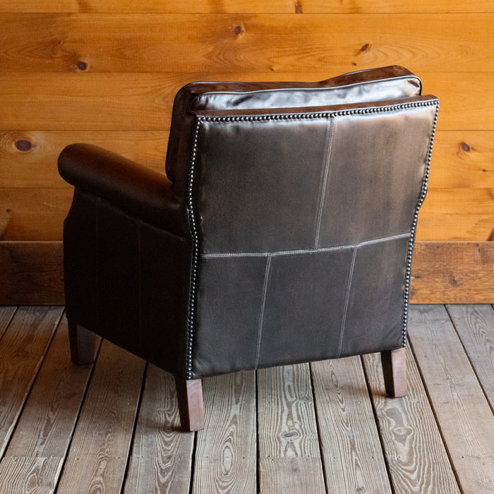 Angled back view of a rustic dark brown leather chair with rolled arms and tapered legs against a wooden wall 