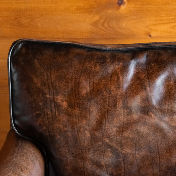 Close-up of the back cushion of a rustic dark brown leather chair against a wooden wall 
