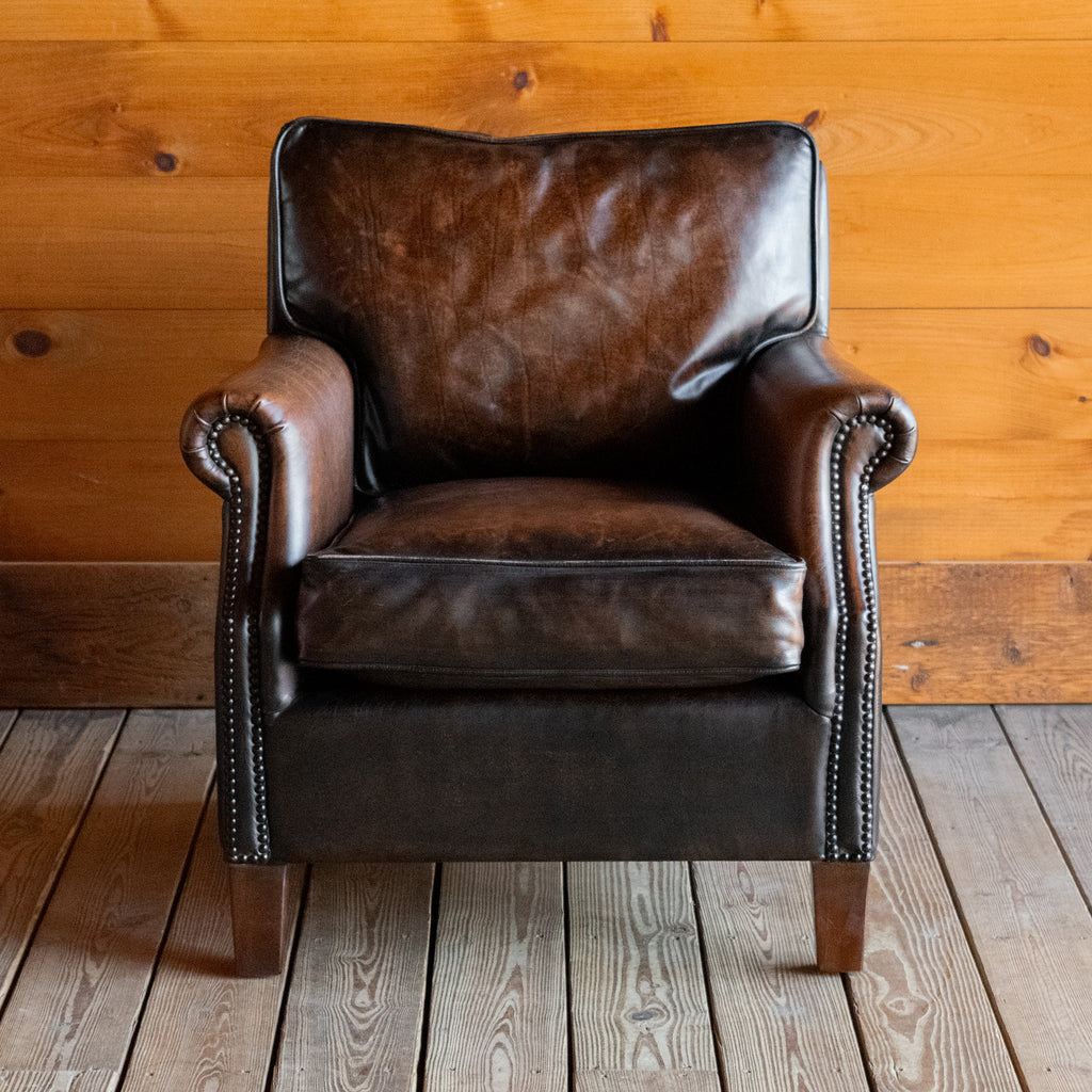 Front view of a rustic dark brown leather chair with rolled arms and tapered legs against a wooden wall 