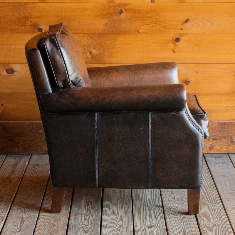 Profile view of a rustic dark brown leather chair with rolled arms and tapered legs against a wooden wall 
