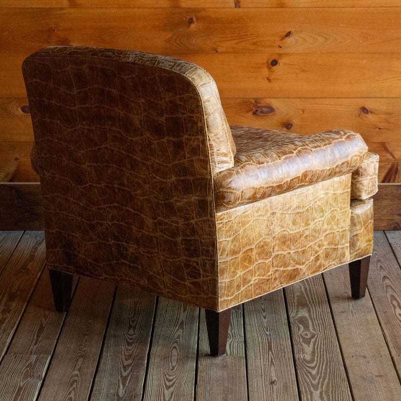 Angled back view of a large, rolled arm club chair upholstered in embossed honey-colored leather with nailhead trim and tapered legs on a wide-plank wooden floor against a rustic wooden wall 
