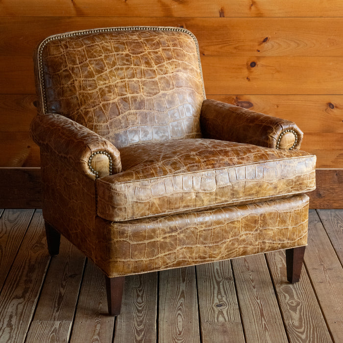 Angled front view of a large, rolled arm club chair upholstered in embossed honey-colored leather with nailhead trim and tapered legs on a wide-plank wooden floor against a rustic wooden wall 