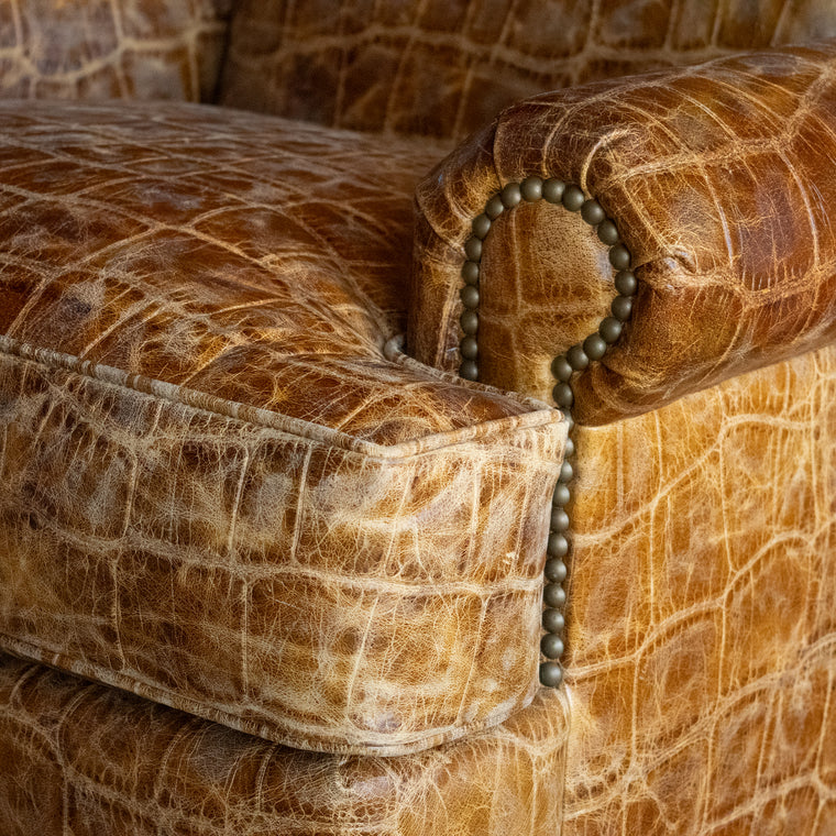 Angled back view of a large, rolled arm club chair upholstered in embossed honey-colored leather with nailhead trim and tapered legs on a wide-plank wooden floor against a rustic wooden wall 