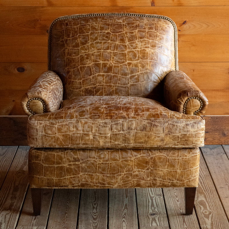 Front view of a large, rolled arm club chair upholstered in embossed honey-colored leather with nailhead trim and tapered legs on a wide-plank wooden floor against a rustic wooden wall 