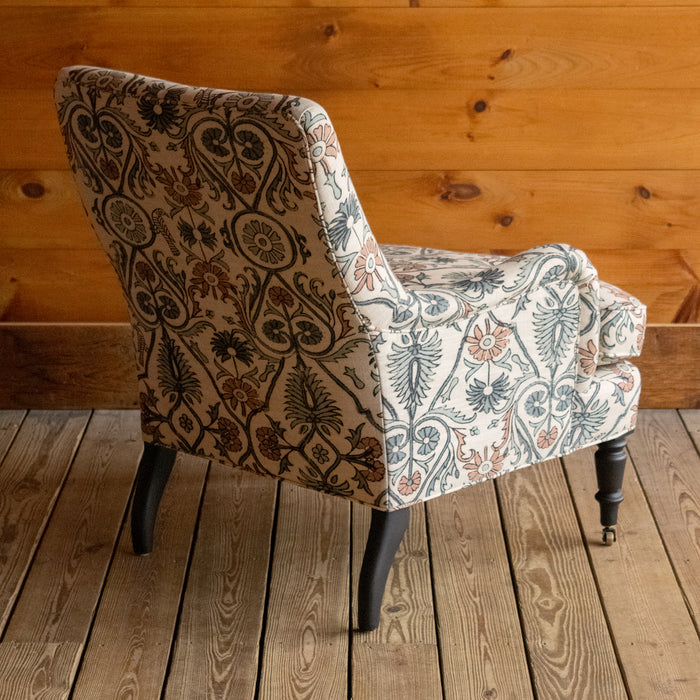 Angled back view of an English rolled arm chair, upholstered in a bird and botanical block print fabric, showcasing its tailored tight back design and flared back legs on a wide-plank wooden floor against a rustic wood-paneled wall