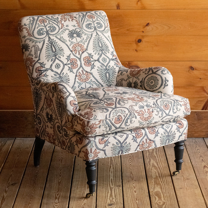 Angled front view of an English rolled arm chair, upholstered in a bird and botanical block print fabric, featuring turned front legs with casters on a wide-plank wooden floor against a rustic wood-paneled wall