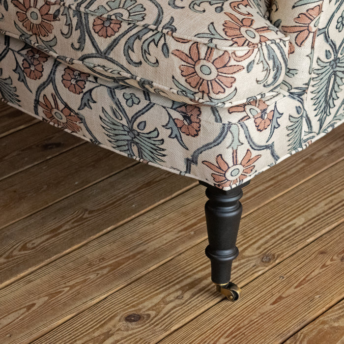 Close-up of a dark-stained turned wooden leg with a caster, supporting an upholstered chair in a bird and botanical block print fabric on a wide-plank wooden floor