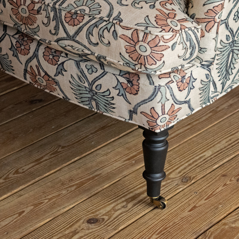 Close-up of a dark-stained turned wooden leg with a caster, supporting an upholstered chair in a bird and botanical block print fabric on a wide-plank wooden floor