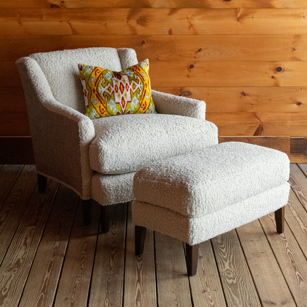Angled front view of a tight back faux shearling chair and ottoman with a vibrant throw pillow on a wide-plank wooden floor against a rustic wooden wall