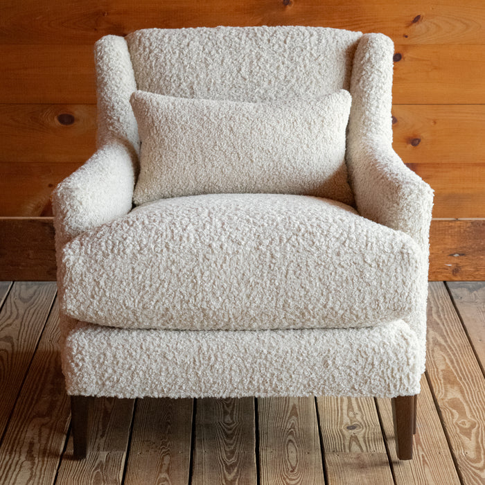 Front view of a tight back faux shearling chair and matching kidney pillow on a wide-plank wooden floor against a rustic wooden wall