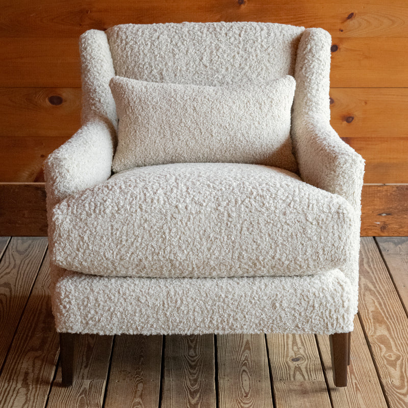 Front view of a tight back faux shearling chair and matching kidney pillow on a wide-plank wooden floor against a rustic wooden wall