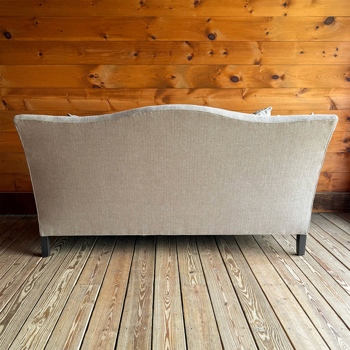 Back view of a bench seat cushion, tight back apartment sofa with flared arms, down throw pillows, and tapered legs on a wooden floor against a wooden wall
