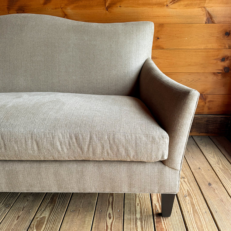 Back view of a bench seat cushion, tight back apartment sofa with flared arms, down throw pillows, and tapered legs on a wooden floor against a wooden wall