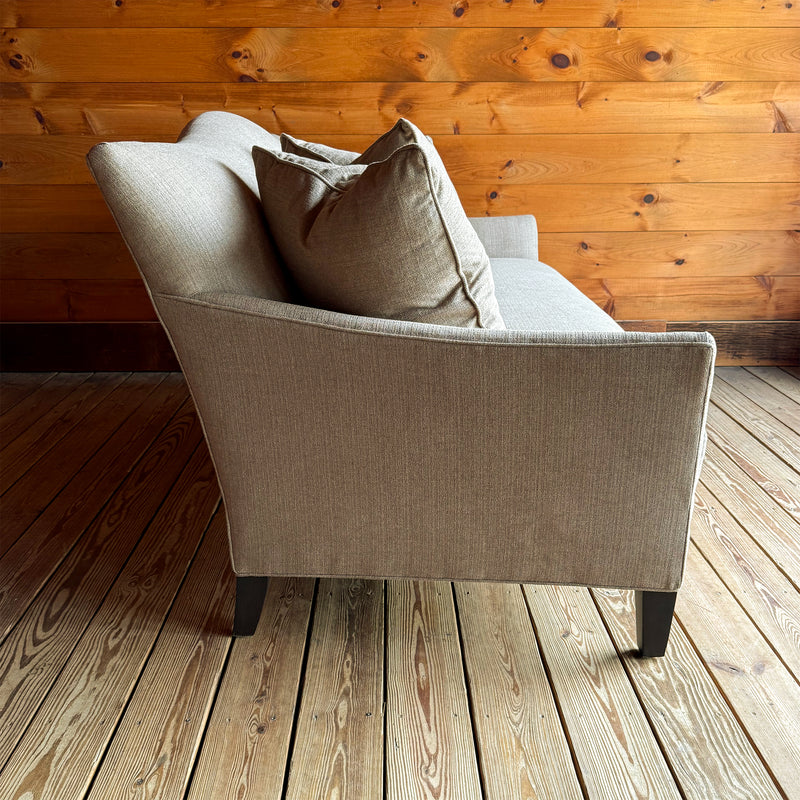 Profile view of a bench seat cushion, tight back apartment sofa with flared arms, down throw pillows, and tapered legs on a wooden floor against a wooden wall
