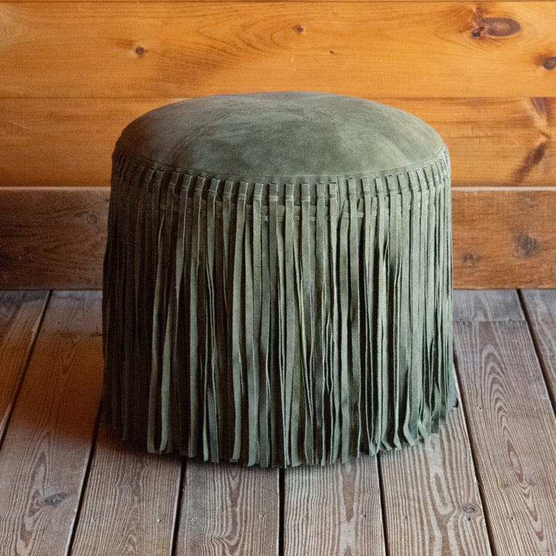 Rustic green suede leather ottoman with fringe on a wooden floor against a wooden wall