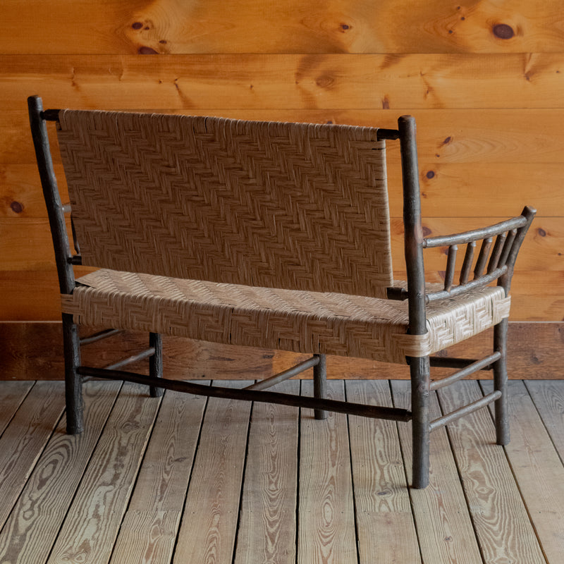 Angled back view of a hickory bench with a paper splint seat and back on a wide-plank wooden floor against a rustic wooden wall