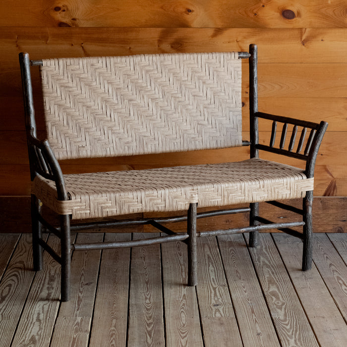 Angled front view of a hickory bench with a paper splint seat and back on a wide-plank wooden floor against a rustic wooden wall