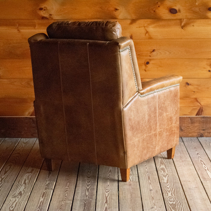 The angled back view of a muted brown leather recliner with nailhead trim against a wooden wall