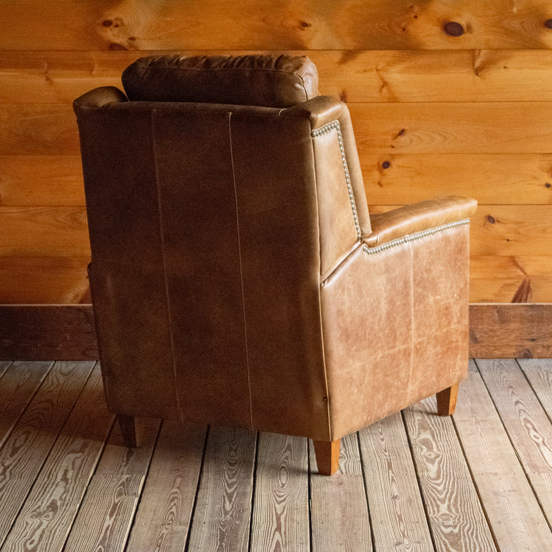 The angled back view of a muted brown leather recliner with nailhead trim against a wooden wall