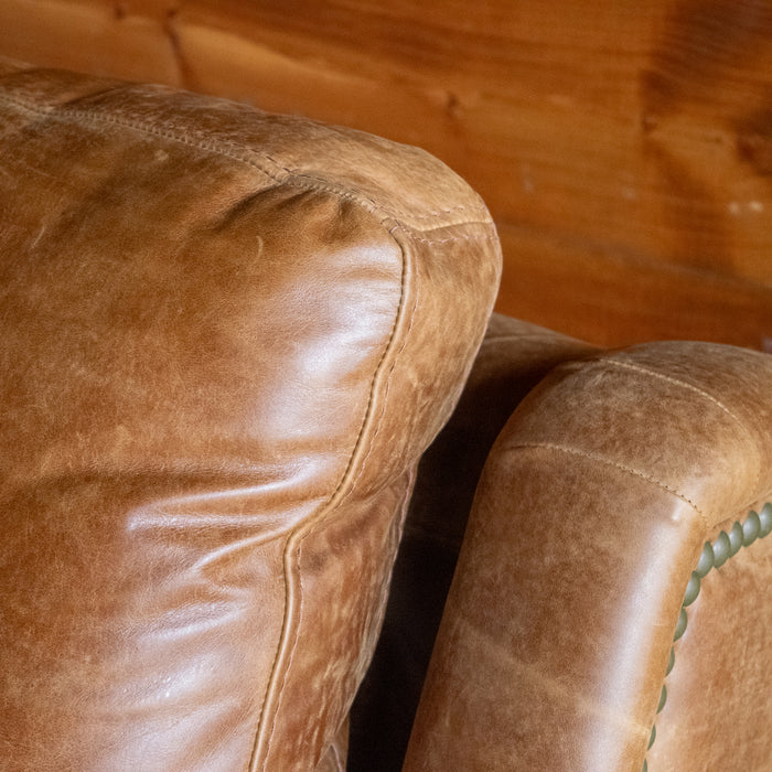 A close-up of the back cushion of a muted brown leather recliner with nailhead trim against a wooden wall